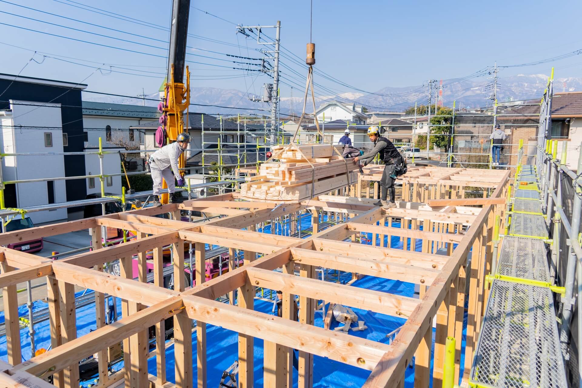 山梨県の住宅現場で、クレーンで吊り上げられた木材を搬入する職人たちの様子。骨組みが整い、背景には山々が広がる。