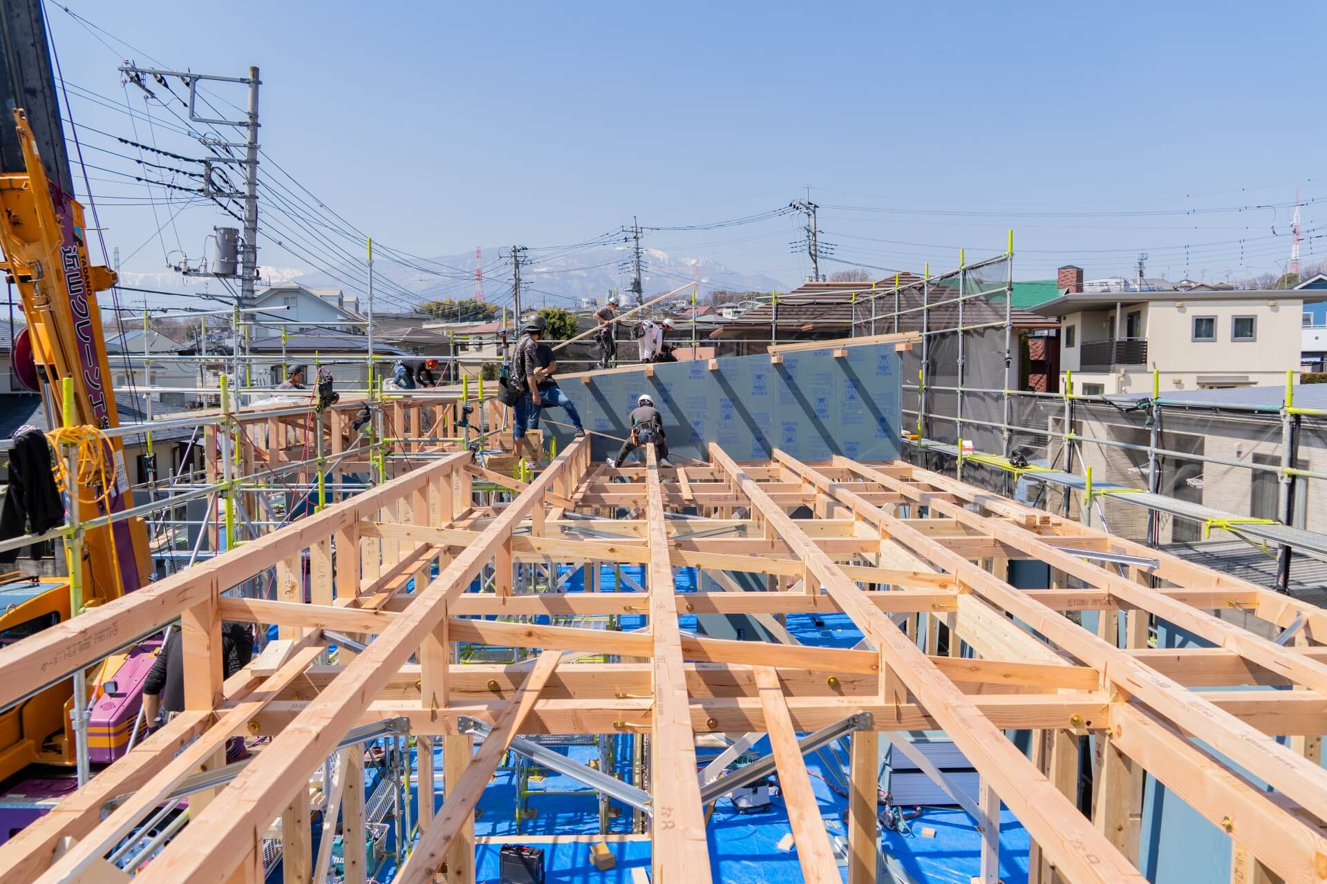 山梨県の住宅現場で、梁の施工が完了し、次の工程へ進む様子。職人たちが足場の上で作業を行い、奥には山々が広がる。