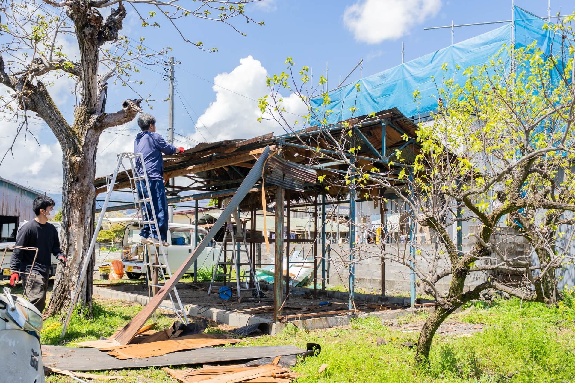 小屋の屋根を脚立に登って外している男性と、その様子を見守るもう一人の男性の姿。小屋は解体作業中で骨組みが露出している。