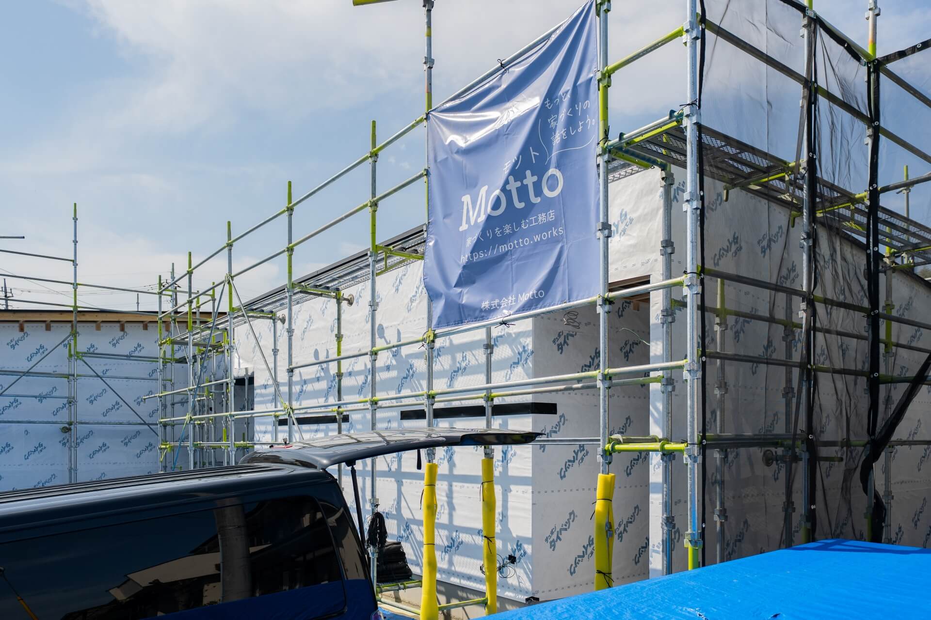 山梨県の住宅建築現場で、外壁下地材が貼られた建物とMottoの足場幕の様子。