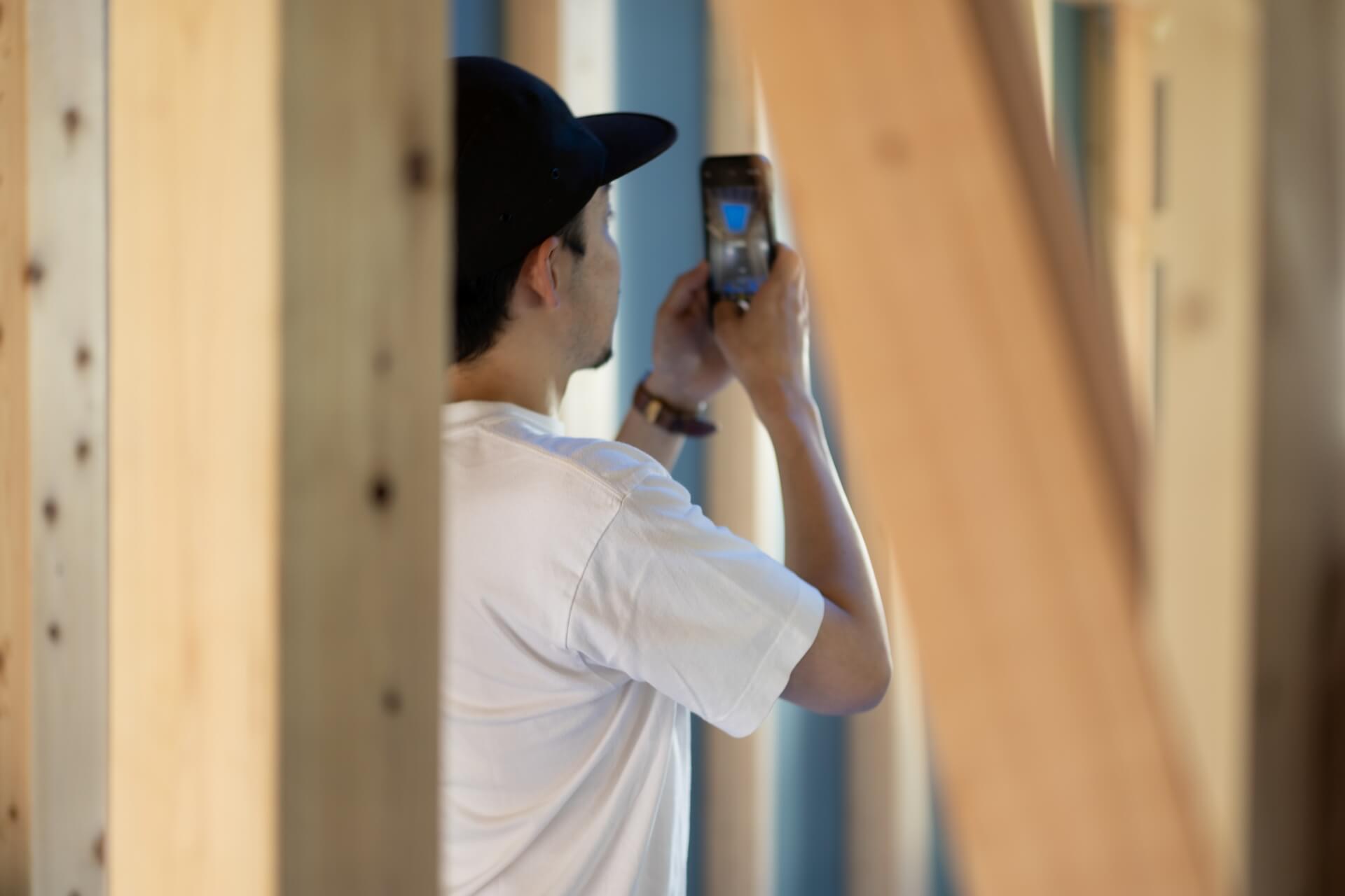 山梨県富士ケ嶺で建築中のヴィラ、スマートフォンを使って撮影しているオーナー。柱の隙間から撮影する姿が映っている。木の質感と柔らかい光が印象的な現場の一コマ。