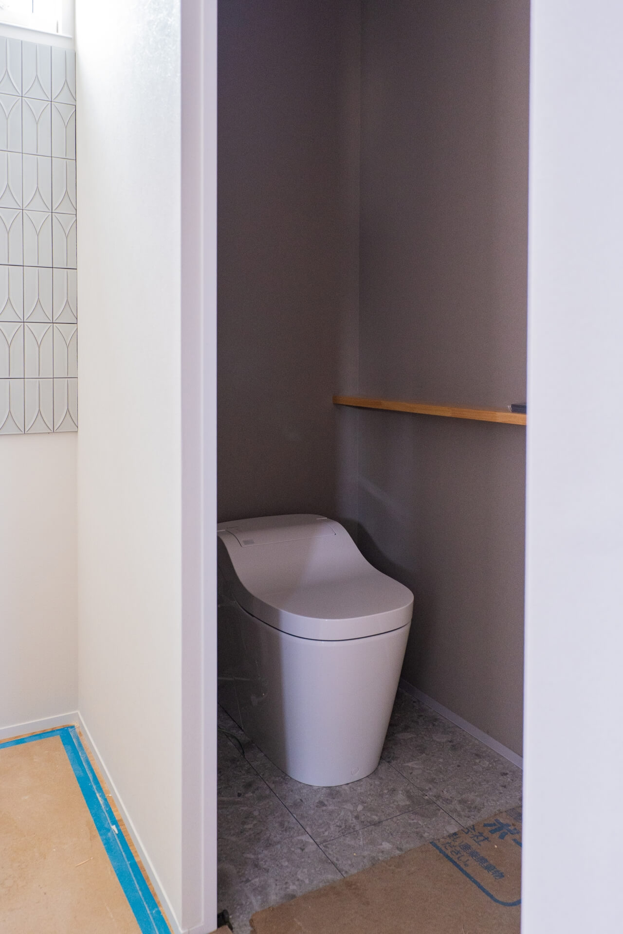 山梨県甲斐市竜王新町の建築現場。トイレはグレーの壁と石目調の床、奥にはタンクレストイレが設置されている。左側の壁には幾何学模様のタイルが見える。