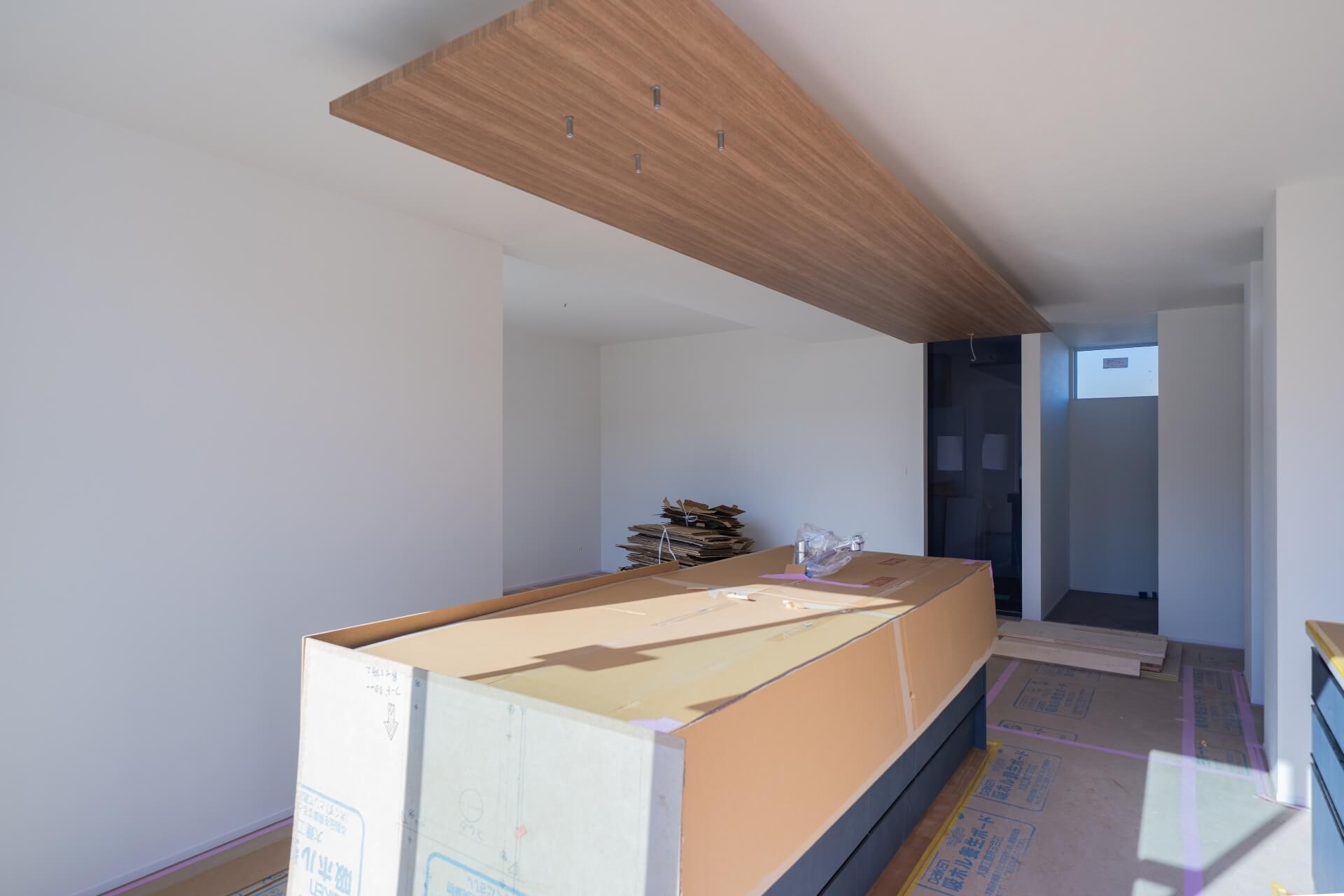 山梨県南アルプス市藤田の現場にて木目の下がり天井と造作キッチンが設けられた住宅内部。施工途中の様子で、空間の中心にキッチンが配置されている。