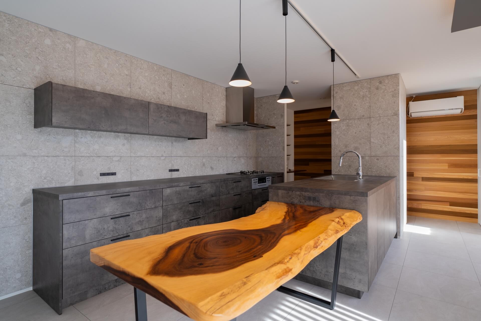 山梨県南アルプス市寺部の新築現場、モルタル調のキッチンと石調タイルに、存在感のある一枚板カウンターを組み合わせた空間。
