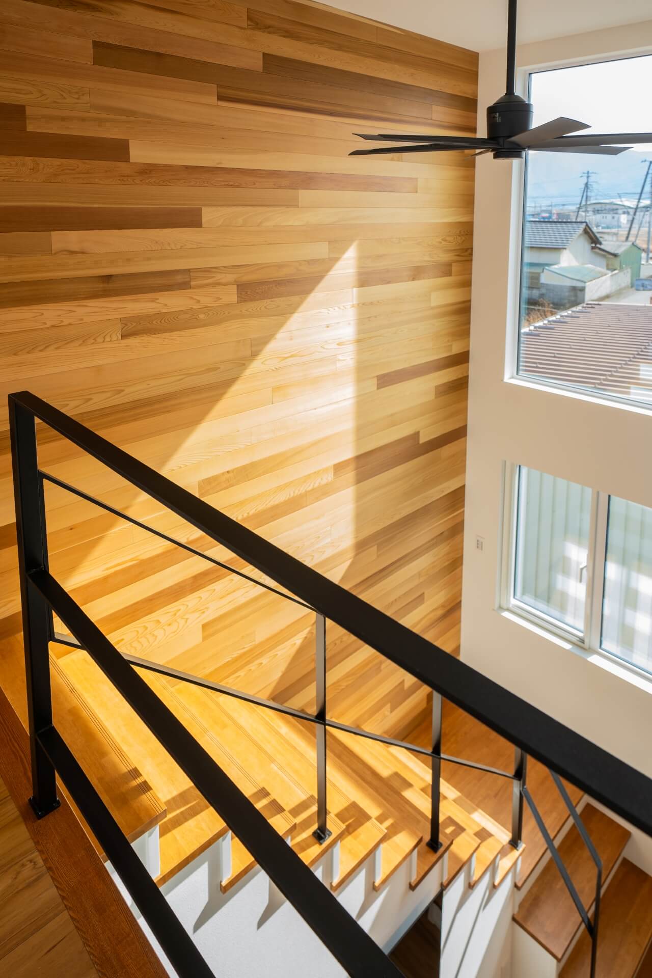 山梨県南アルプス市寺部の新築現場、吹抜けの大きな窓から差し込む光が、木張りの壁に陰影をつくる空間。