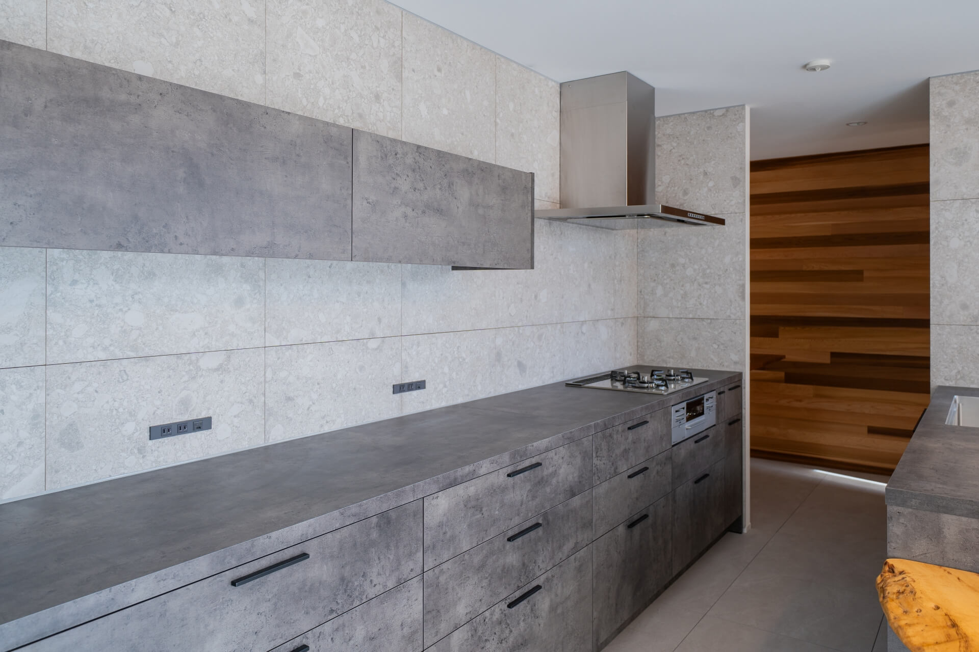 山梨県南アルプス市寺部の新築現場、石調タイルとフラットなグレーのキッチン収納が連続する、ミニマルな壁面デザイン。