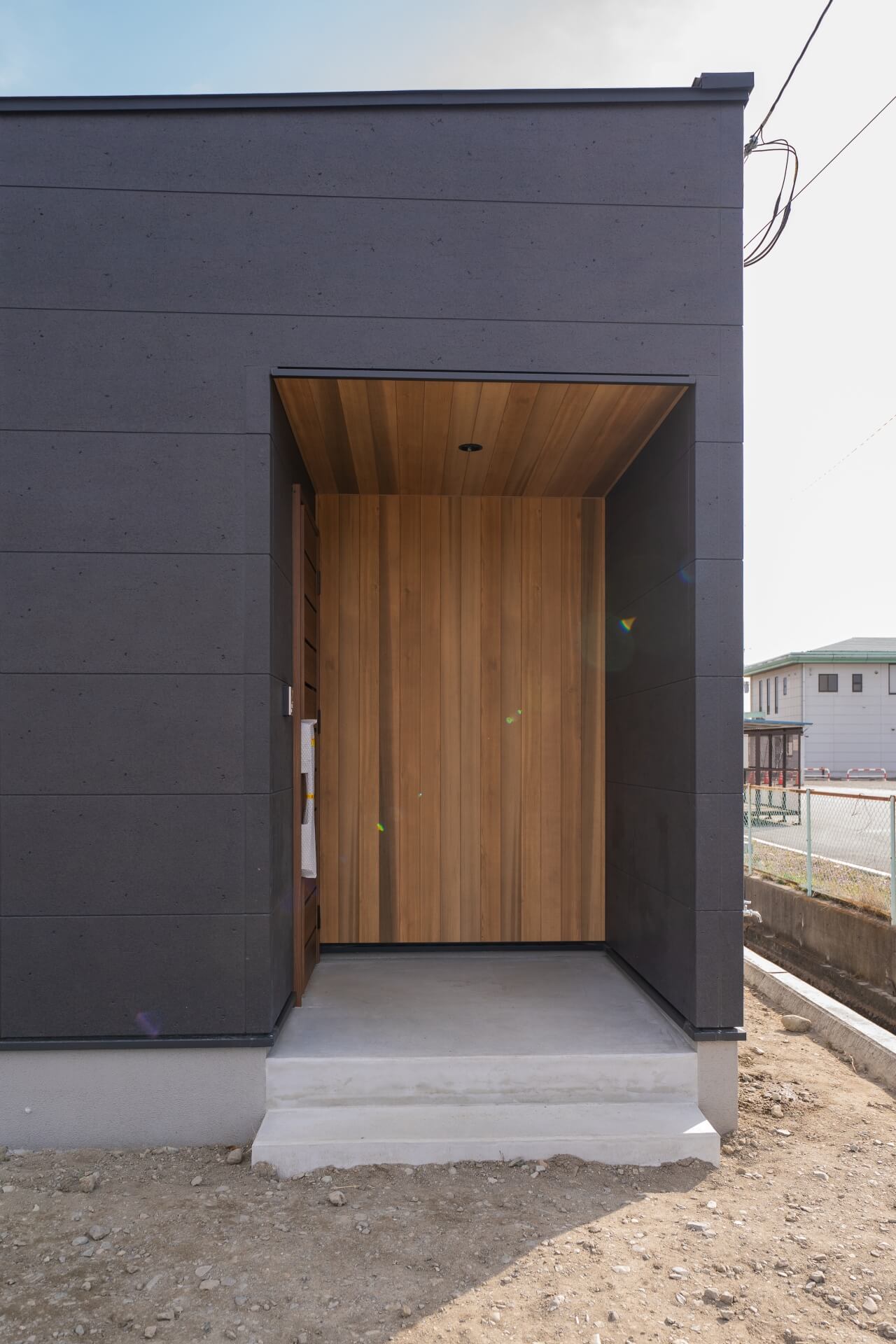山梨県南アルプス市藤田の黒い外壁に木張りの玄関ポーチが切り取られた平屋住宅の外観。陰影が印象的なアプローチ空間。